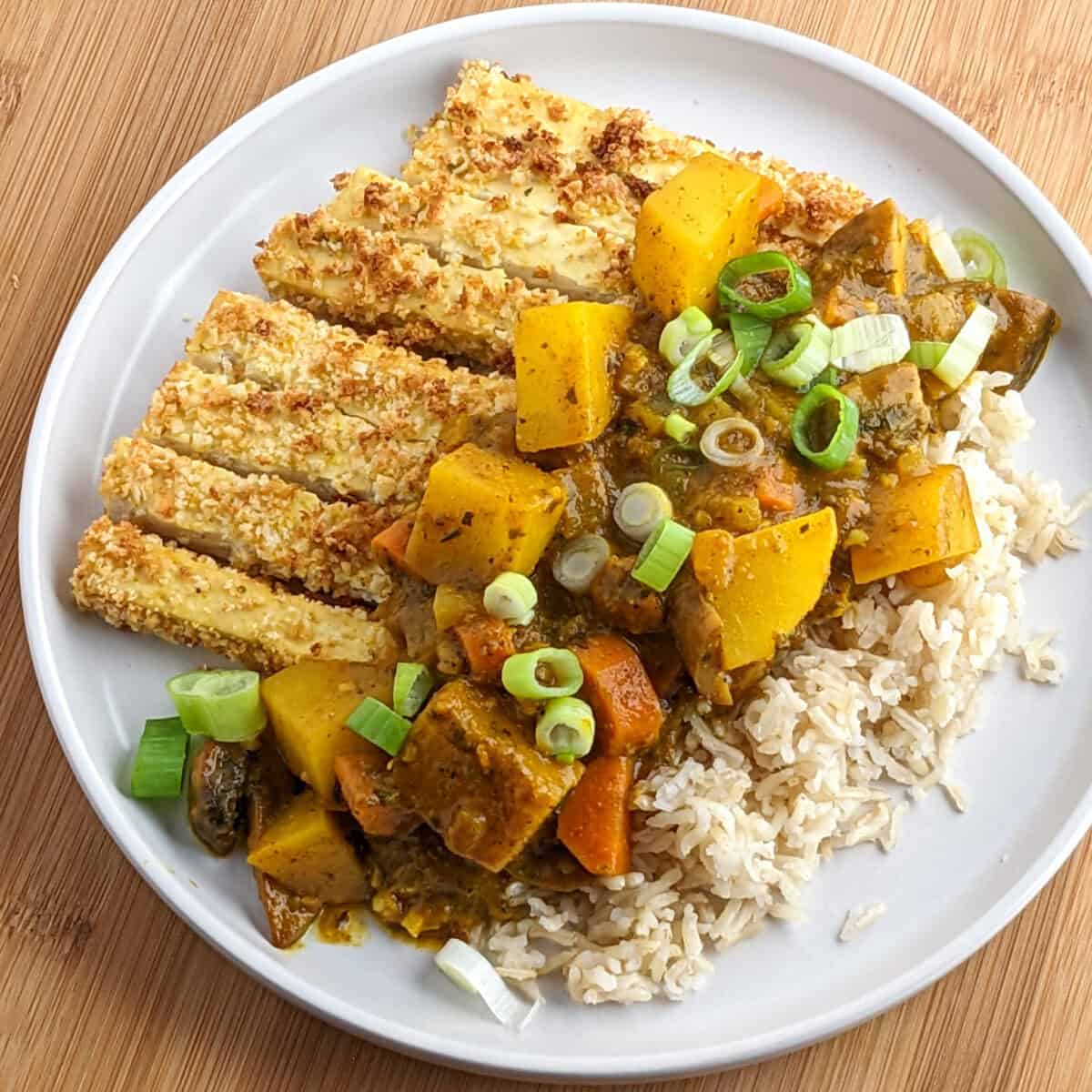 Breaded sliced tofu and brown rice topped with curried potatoes, carrots and onions on a white round plate.